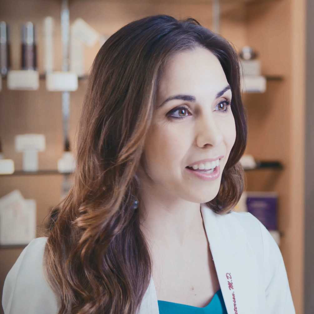 Smiling doctor in a white coat standing in front of a shelf of skincare products at a medical aesthetic clinic in Texas.