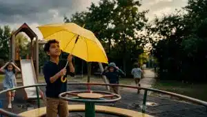 Child holding a yellow umbrella on a playground during light rain.